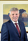 Mann i dress og slips poserer foran et innrammet maleri av et hus ved vann.